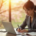 A woman in a gray blazer works at a desk with a laptop and open notebook, writing with a pen as she uses Smart PDF Merging software. Sunlight streams through large windows with a blurred view of trees and pink blossoms outside. | LESSINVEST