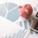A pink piggy bank, calculator, and various financial charts and graphs are arranged on a white surface, representing financial planning or budgeting. | LESSINVEST