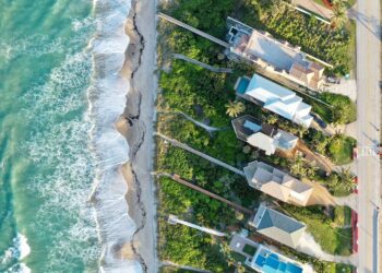 Aerial view of ocean waves crashing onto a sandy beach beside a row of houses with gardens and pools, separated from the shoreline by lush greenery and a narrow road. | LESSINVEST