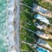 Aerial view of ocean waves crashing onto a sandy beach beside a row of houses with gardens and pools, separated from the shoreline by lush greenery and a narrow road. | LESSINVEST
