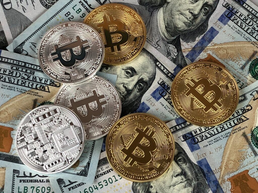 Gold and silver Bitcoin coins are scattered on top of several U.S. $100 bills featuring Benjamin Franklin’s portrait, representing the concept of cryptocurrency and traditional currency together. | LESSINVEST