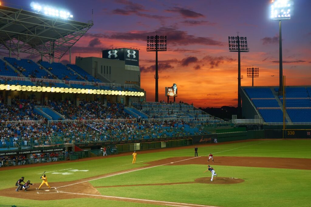 A baseball game in progress at a stadium during sunset, with players on the field, spectators in the stands, and a dramatic orange and purple sky in the background. | LESSINVEST