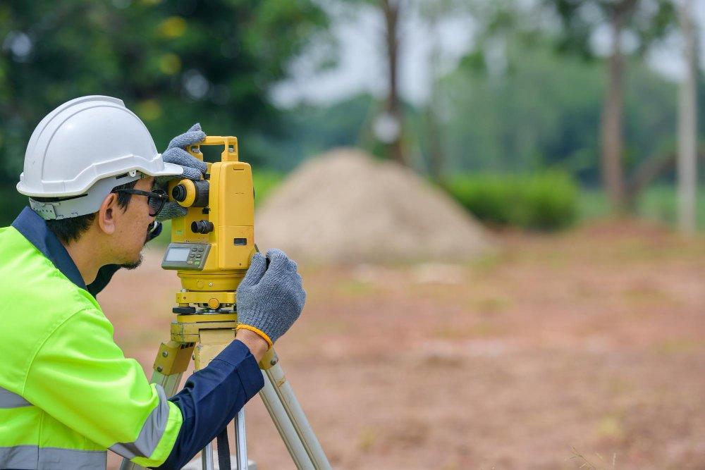 A surveyor in a white hard hat, safety glasses, gloves, and a high-visibility jacket uses a yellow theodolite on a tripod to conduct Topographical Surveys at an outdoor construction site with trees and soil in the background. | LESSINVEST
