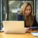 A woman sits at a desk with a laptop, looking at a tablet. Surrounded by papers, a cup, and a phone, she appears to be working in a modern office—perhaps finding ways to help your budget behind sleek glass doors. | LESSINVEST