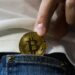 A person places a gold-colored Bitcoin coin into the front pocket of blue jeans, with a white shirt visible in the background. | LESSINVEST