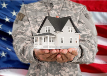 A person in military uniform holds a model house in their hands, symbolizing homeownership made possible through a VA Loan, with a blurred American flag in the background. | LESSINVEST