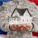 A person in military uniform holds a model house in their hands, symbolizing homeownership made possible through a VA Loan, with a blurred American flag in the background. | LESSINVEST