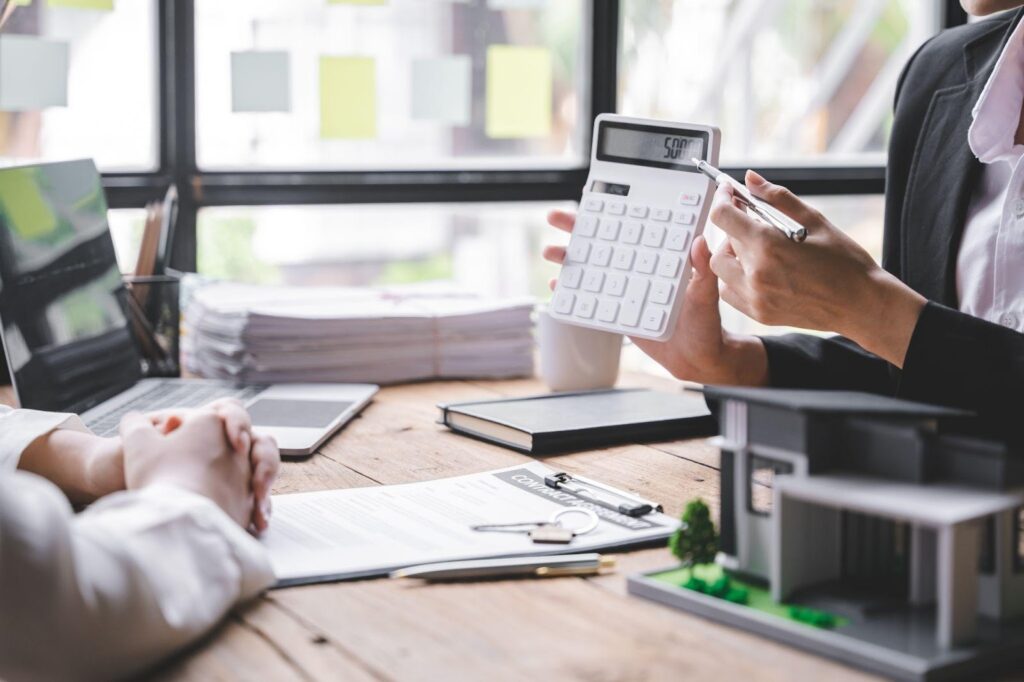 Two people sit at a desk with documents, a calculator, and a model house. One person points at the calculator display while discussing with the other, suggesting a financial or real estate consultation. | LESSINVEST