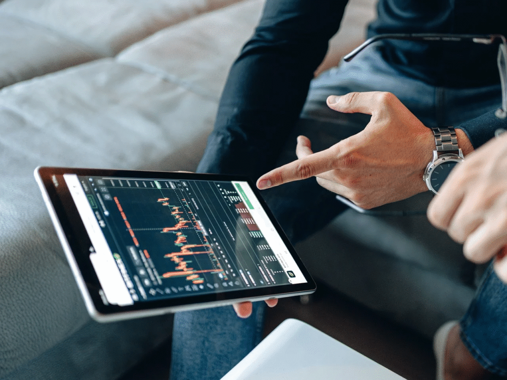 Two people review a tablet displaying a candlestick stock chart with green and red bars. One person points at the screen, and both are seated on a couch. | LESSINVEST