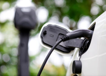 A close-up of an electric vehicle being charged, showing the charging cable plugged into the car’s port. A blurred charging station and green foliage are visible in the background. | LESSINVEST