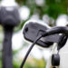 A close-up of an electric vehicle being charged, showing the charging cable plugged into the car’s port. A blurred charging station and green foliage are visible in the background. | LESSINVEST