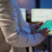 A person in a white blazer uses a tablet displaying financial charts and data, standing in an office with blurred computer screens in the background. | LESSINVEST