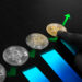 A hand points to a row of silver and gold bitcoin coins placed along a green upward arrow, with blue bar graphs rising underneath, symbolizing growth in cryptocurrency value. | LESSINVEST