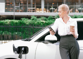 A woman in professional attire stands beside a white electric car being charged, holding a coffee cup and looking at her smartphone. Greenery and a modern building are visible in the background. | LESSINVEST