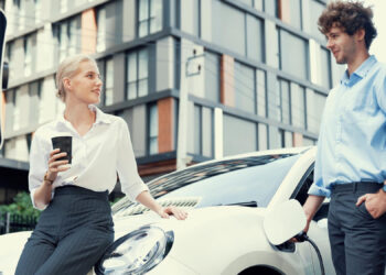 A woman holding a coffee cup and a man stand by a white electric car at a charging station, smiling at each other, with a modern building in the background. | LESSINVEST