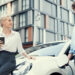 A woman holding a coffee cup and a man stand by a white electric car at a charging station, smiling at each other, with a modern building in the background. | LESSINVEST