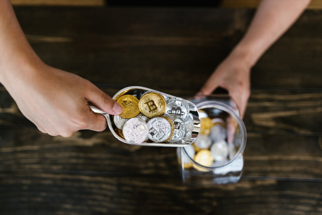 A person uses a metal scoop to transfer various cryptocurrency coins, including Bitcoin and Ethereum, from a glass jar onto a dark wooden surface. | LESSINVEST