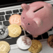 A pink piggy bank and several gold, silver, and copper Bitcoin coins are placed on the keyboard of a laptop. | LESSINVEST