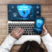 A person using a laptop and smartphone displaying a cyber security graphic with a shield and padlocks, viewed from above on a wooden desk. | LESSINVEST
