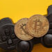Two gold Bitcoin coins are placed on the buttons of a black video game controller, with a bright yellow background. | LESSINVEST