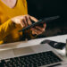 A person in a yellow top uses a tablet at a desk with a laptop, a jar of coins, a calculator, and a remote control. | LESSINVEST
