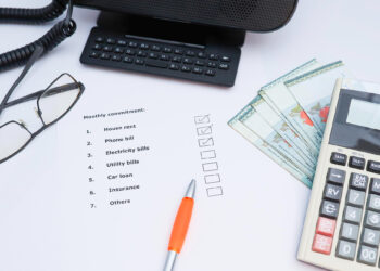 A checklist titled "Monthly commitment" on paper with items like house rent and phone bill, a pen, calculator, glasses, phone, and several 100 currency notes on a white desk. | LESSINVEST