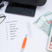 A checklist titled "Monthly commitment" on paper with items like house rent and phone bill, a pen, calculator, glasses, phone, and several 100 currency notes on a white desk. | LESSINVEST