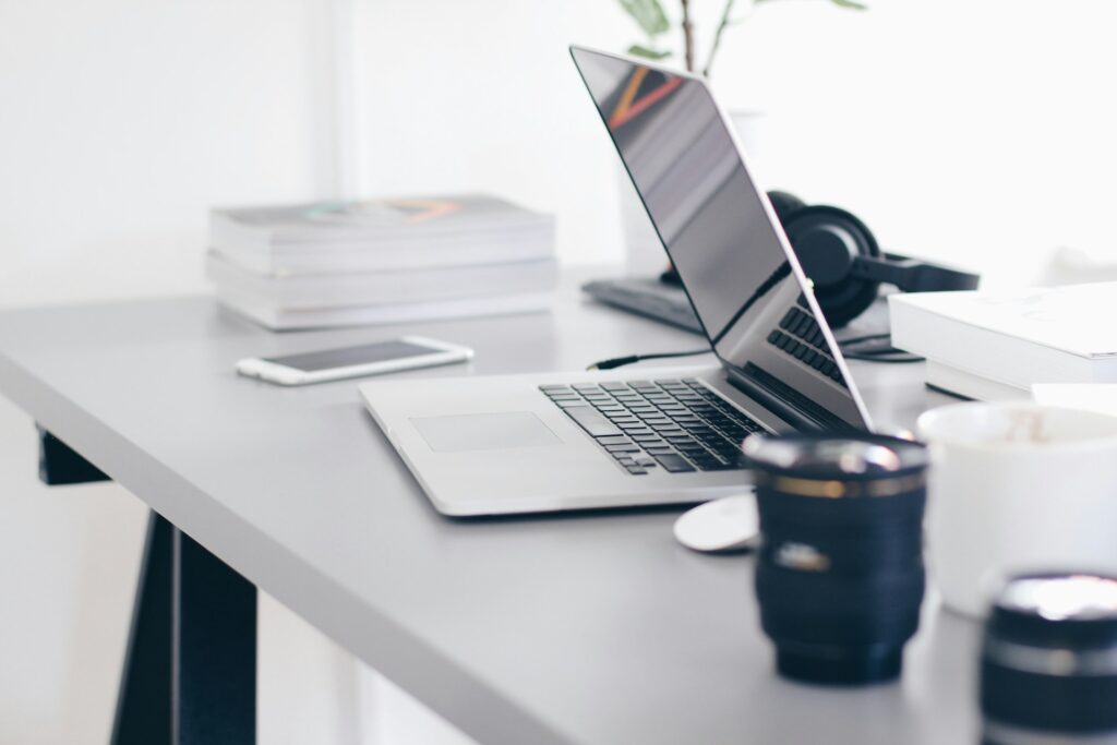 A sleek, modern desk with an open laptop, smartphone, camera lens, headphones, stacked magazines, and a white mug in a bright, minimalist workspace. | LESSINVEST