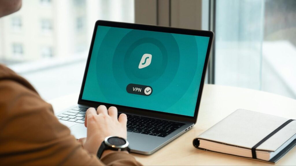 A person using a laptop with a VPN application open on the screen, sitting at a round table near a window, with a closed notebook and pen beside the laptop. | LESSINVEST