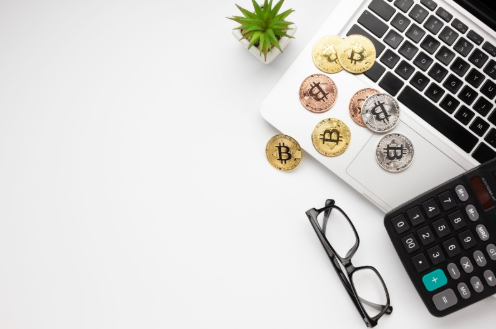 A laptop with several Bitcoin coins, a calculator, eyeglasses, and a small potted plant on a white desk, symbolizing crypto investing, finance, and technology. | LESSINVEST