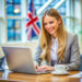 A woman in business attire smiles while working on a laptop at a desk with a cup of coffee, a plant, and a tablet; a British flag is visible in the background. | LESSINVEST