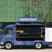 A blue food truck parked in front of a modern glass building, with a menu displayed on its side and "FOODTRUCK MOCKUP" sign above. The service window is open, and a tree with sparse branches is nearby. | LESSINVEST