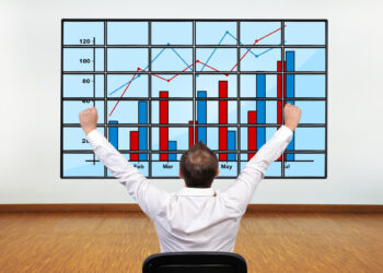 A man in a white shirt sits facing a large screen displaying bar and line graphs, raising his arms in celebration as the charts show positive data trends. | LESSINVEST