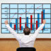 A man in a white shirt sits facing a large screen displaying bar and line graphs, raising his arms in celebration as the charts show positive data trends. | LESSINVEST
