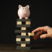 A hand removes a block from a stack of wooden blocks with a pink piggy bank balanced on top, symbolizing financial risk or instability, against a dark background. | LESSINVEST