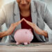 A person in a blazer forms a roof shape with their hands over a pink piggy bank sitting on a table with dollar bills, symbolizing financial protection and savings. | LESSINVEST