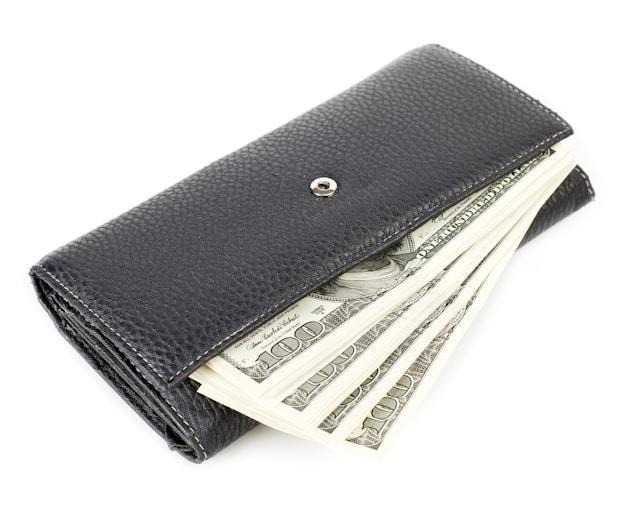 A black leather wallet filled with several U.S. hundred dollar bills sticking out on a white background. | LESSINVEST