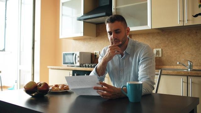 A man sits at a kitchen table, looking thoughtfully at a piece of paper in his hand. A blue mug, a bowl of fruit, and a plate of pastries are on the table. The kitchen is bright and modern. | LESSINVEST