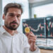 A man in a white shirt sits at a desk holding a gold Bitcoin coin, with two computer monitors behind him displaying cryptocurrency price charts. | LESSINVEST