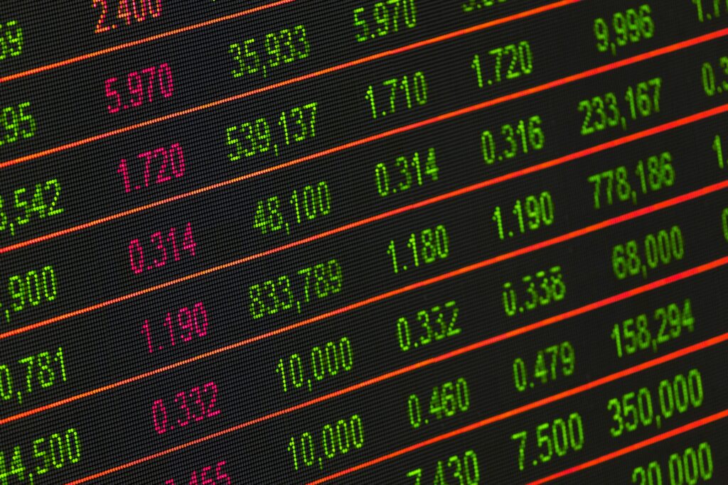 A digital display showing rows of financial data with numbers in red and green, representing stock market prices, volumes, and changes. | LESSINVEST