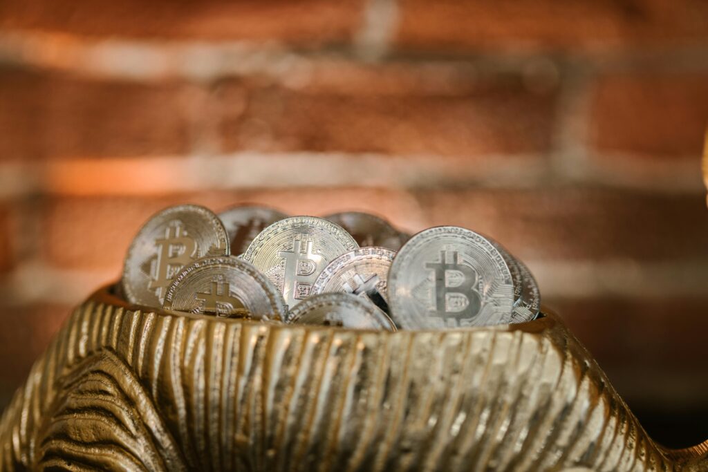 A close-up of several silver-colored Bitcoin tokens placed in a textured, gold-colored dish, with a blurred brick wall background. | LESSINVEST