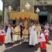A religious procession outside a church, featuring clergy in white and gold robes under a decorative canopy, with altar servers in red and white garments and a crowd of onlookers in the background. | LESSINVEST
