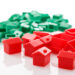 A group of small red and green plastic Monopoly houses and hotels scattered on a white surface. The red pieces are in the foreground, and the green pieces are in the background. | LESSINVEST