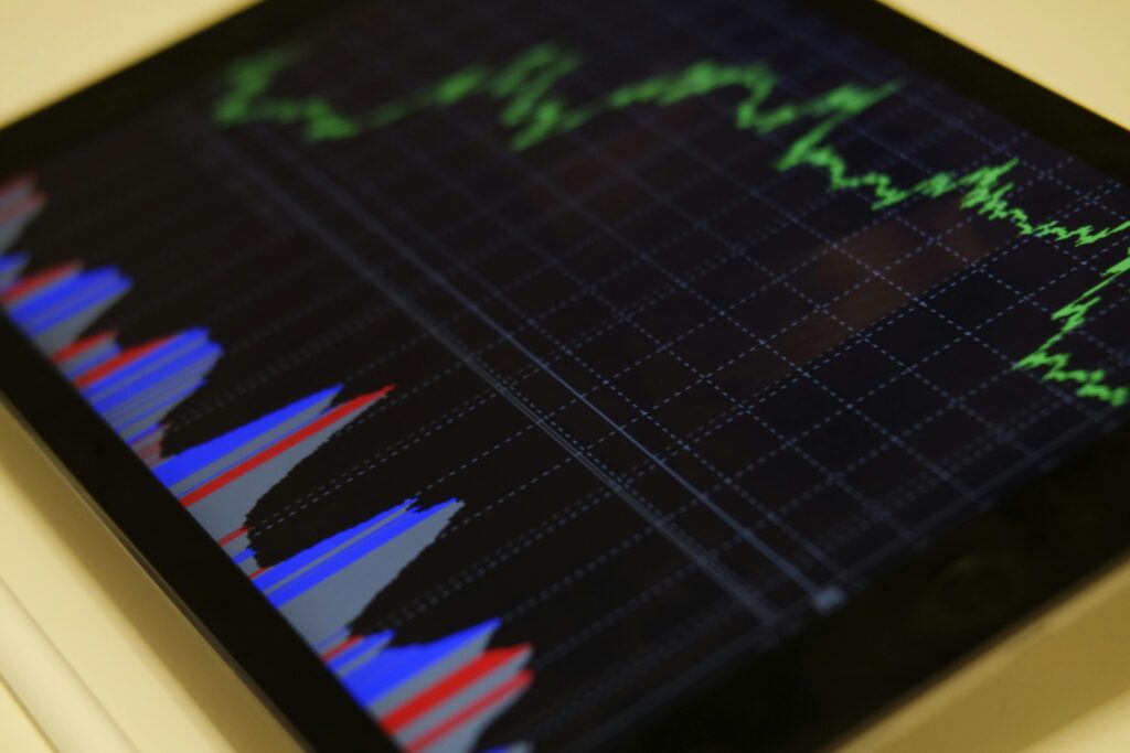 A digital tablet displays a financial chart with green line graphs and blue and red bar graphs, indicating market trends and data analysis. | LESSINVEST