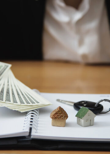 A person in a suit holds out dollar bills with one hand and points with the other at two small house models and a car key placed on an open notebook on a desk. | LESSINVEST