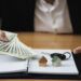 A person in a suit holds out dollar bills with one hand and points with the other at two small house models and a car key placed on an open notebook on a desk. | LESSINVEST