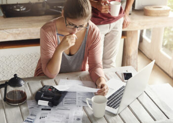 A woman sits at a table with bills, a calculator, and a laptop, looking concerned as she reviews documents. A coffee pot and two mugs are on the table, and another person stands nearby holding a mug. | LESSINVEST