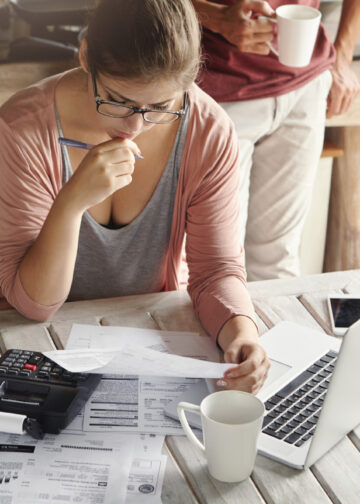 A woman sits at a table with bills, a calculator, and a laptop, looking concerned as she reviews documents. A coffee pot and two mugs are on the table, and another person stands nearby holding a mug. | LESSINVEST