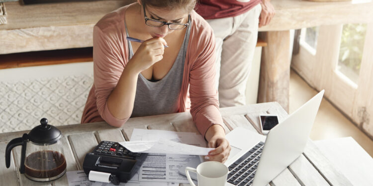 A woman sits at a table with bills, a calculator, and a laptop, looking concerned as she reviews documents. A coffee pot and two mugs are on the table, and another person stands nearby holding a mug. | LESSINVEST