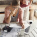 A woman sits at a table with bills, a calculator, and a laptop, looking concerned as she reviews documents. A coffee pot and two mugs are on the table, and another person stands nearby holding a mug. | LESSINVEST